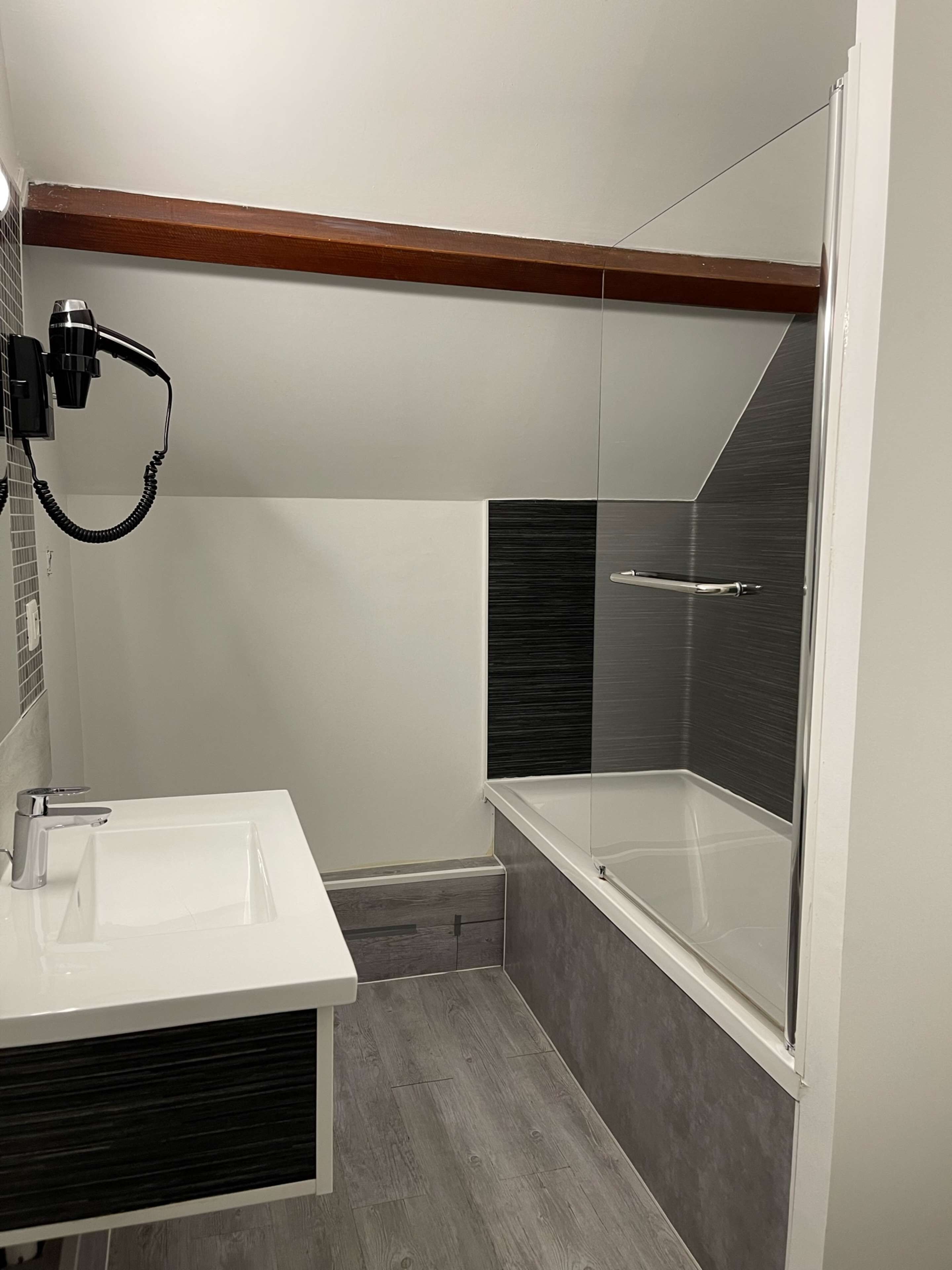 Un baño moderno con un lavabo blanco, cabina de ducha con mampara de vidrio y un travesaño de madera en el techo.