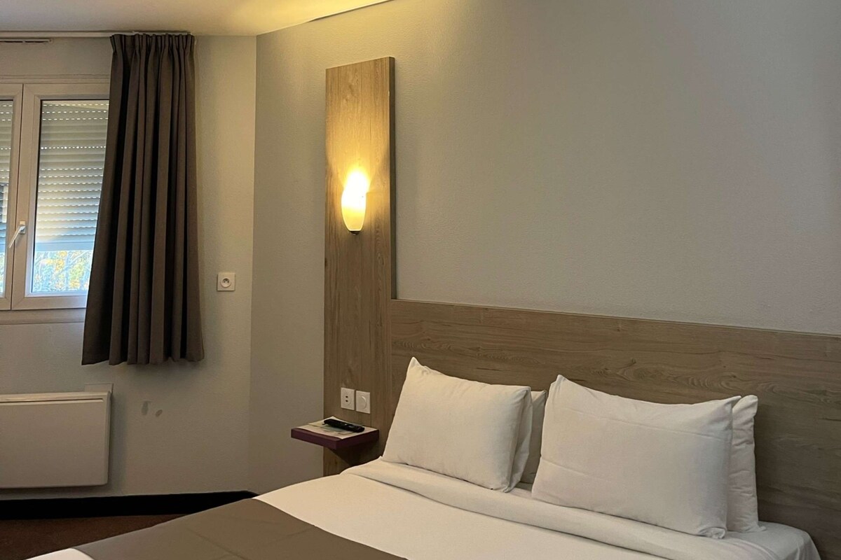 A hotel room with a bed, a nightstand, a wall-mounted light, and a window with curtains.