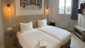 A double bed with white linens and a folded towel, a nightstand, a wall-mounted lamp, and a window with curtains. - Kyriad Saint Fargeau Ponthierry Apollonia (Saint-Fargeau-Ponthierry)
