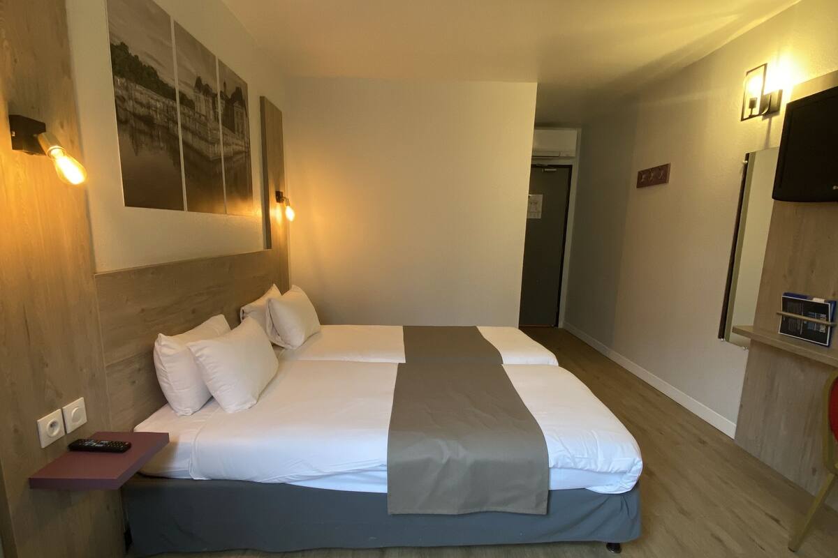 A double bed with a grey blanket, white pillows, and a wooden headboard. There is a wall-mounted TV and a small shelf with a radio. The room has wooden flooring and light-colored walls.