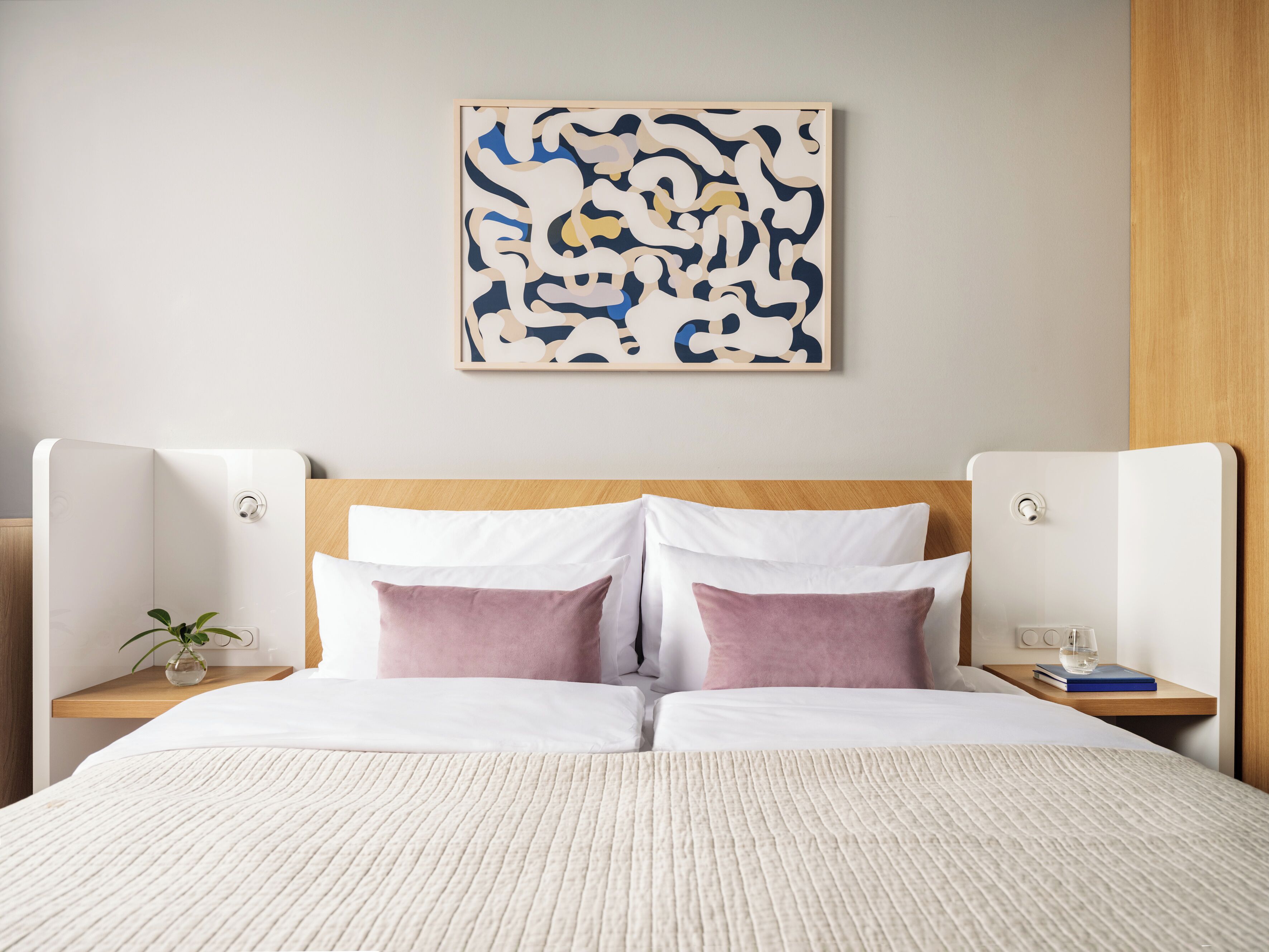 Een net opgemaakt tweepersoonsbed met witte en roze beddengoed, een houten hoofdbord en een lijst met abstracte kunst aan de muur.