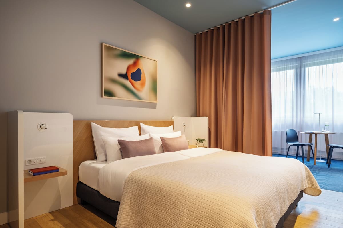 Quarto de hotel com uma cama grande, uma mesa de cabeceira com um livro, uma obra de arte na parede e vista para o exterior através de uma janela com cortinas.