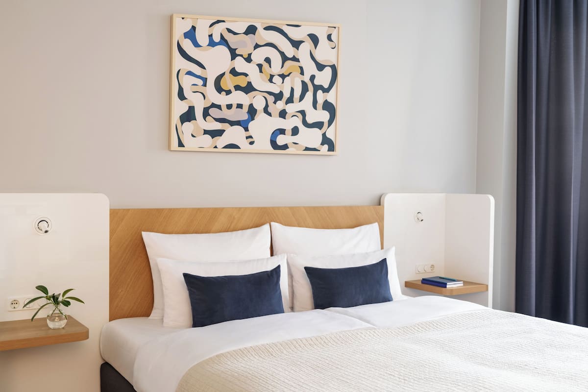 Uma cama bem arrumada, com almofadas brancas e azul escuras, uma cabeceira de madeira e uma mesa de cabeceira com um livro e uma pequena planta.