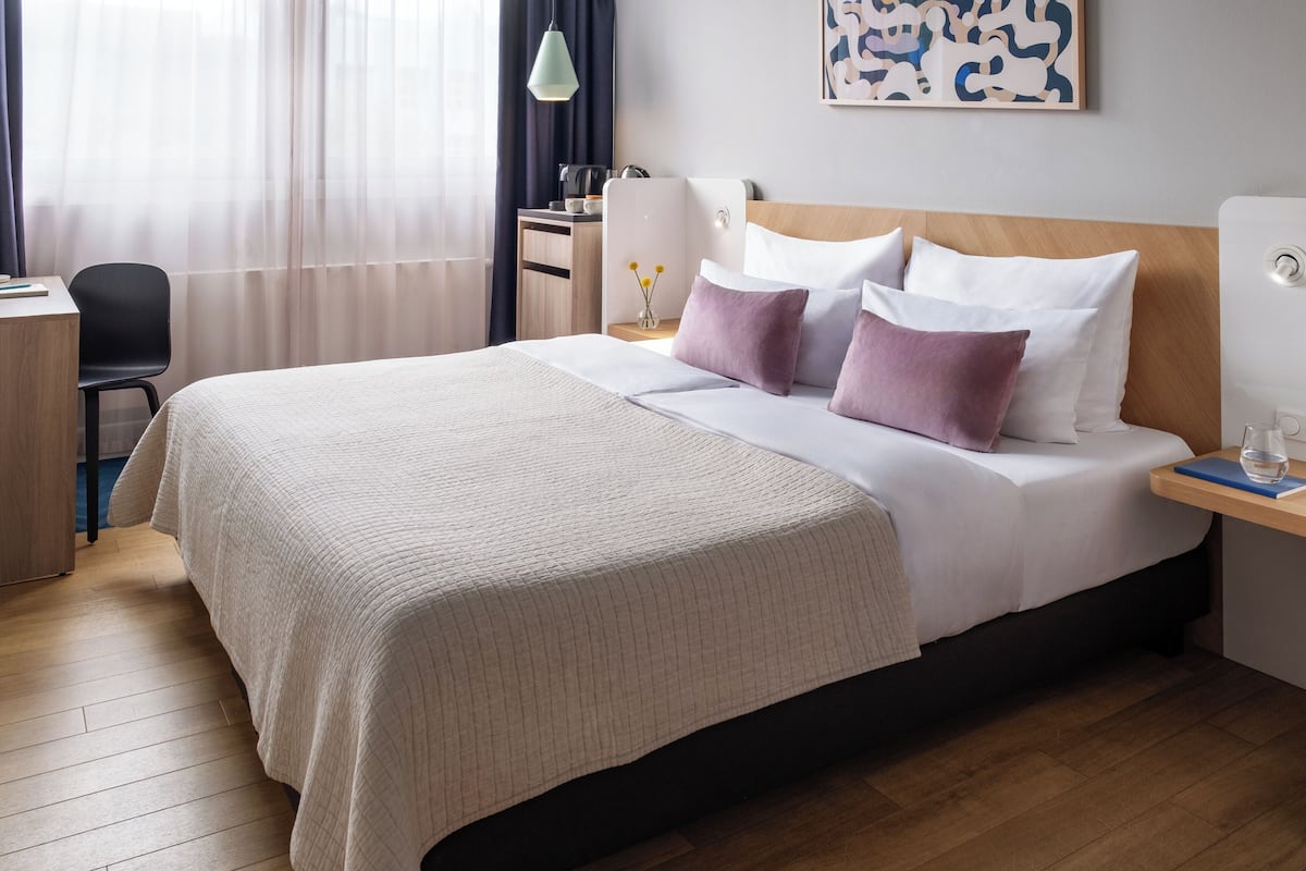 Cama bem arrumada com colcha branca e bege, mesa de cabeceira com um copo de água e escrivaninha com abajur.