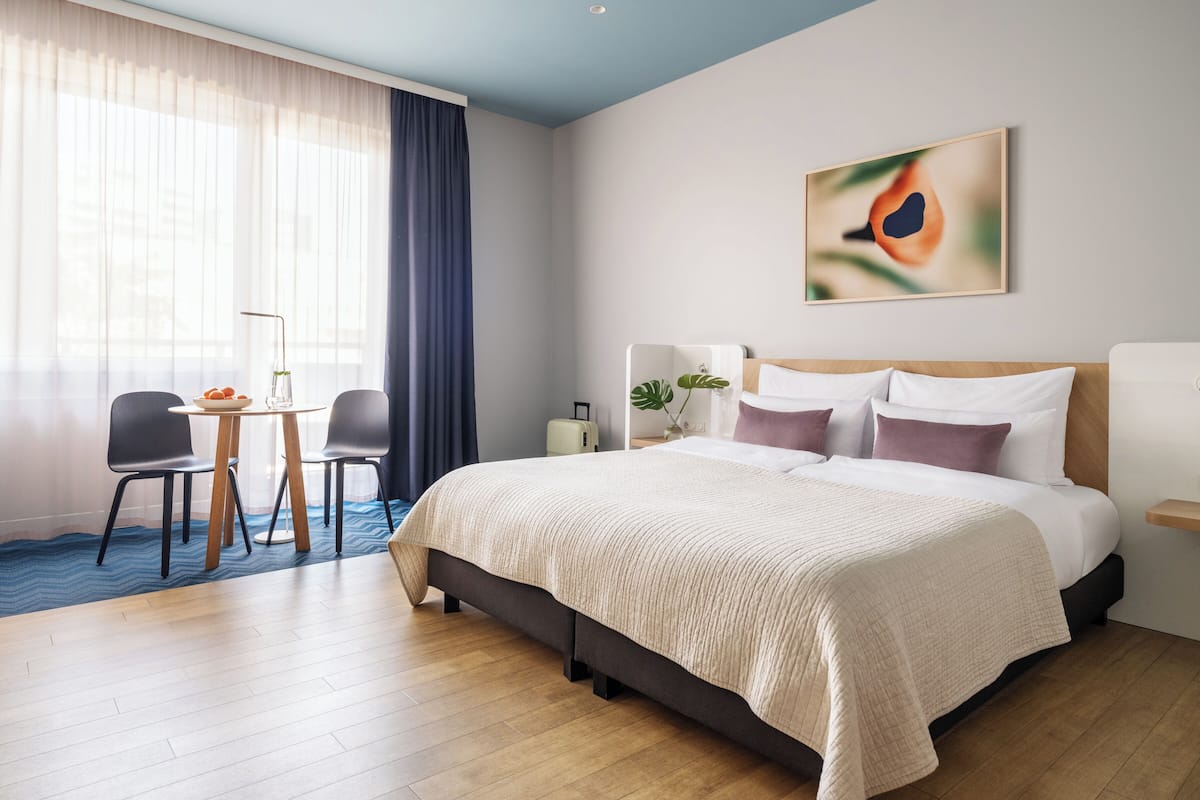 Quarto de hotel moderno com uma cama grande, uma mesa com cadeira, uma mesinha com uma tigela de frutas e uma janela com cortinas leves.