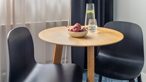 A round wooden table with a bowl of fruit and a glass of water, flanked by two black chairs, in a room with a window and curtains. - Numa Berlin Kudamm (Berlin)