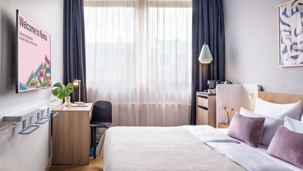 A hotel room with a bed, desk, chair, and a wall-mounted TV displaying a welcome message. - Numa Berlin Kudamm (Berlin)