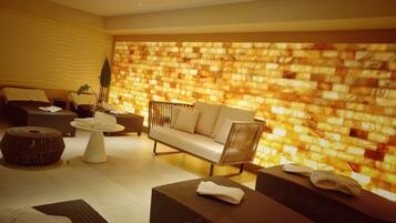 A modern spa room with a textured wall, a sofa, two massage tables, and a small round table with towels.