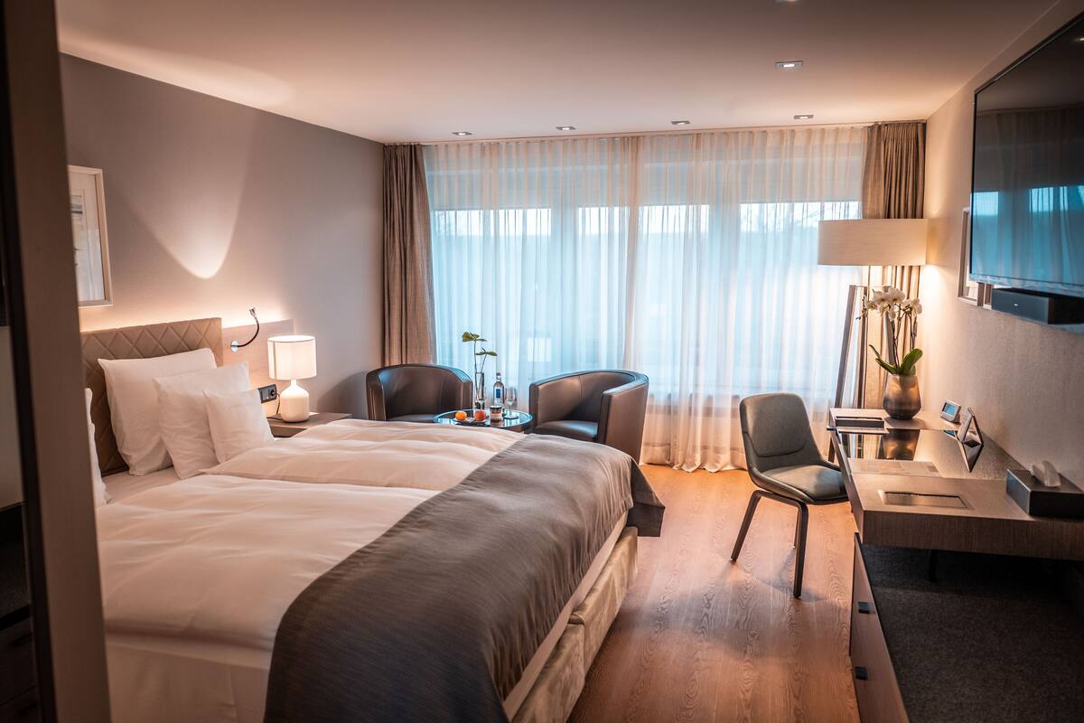 Et hotelværelse med en stor seng, et skrivebord med stol, et fjernsyn og et vindue med let gardin.