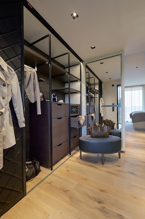 A modern hotel room with a walk-in closet, a round ottoman, and a large bed.