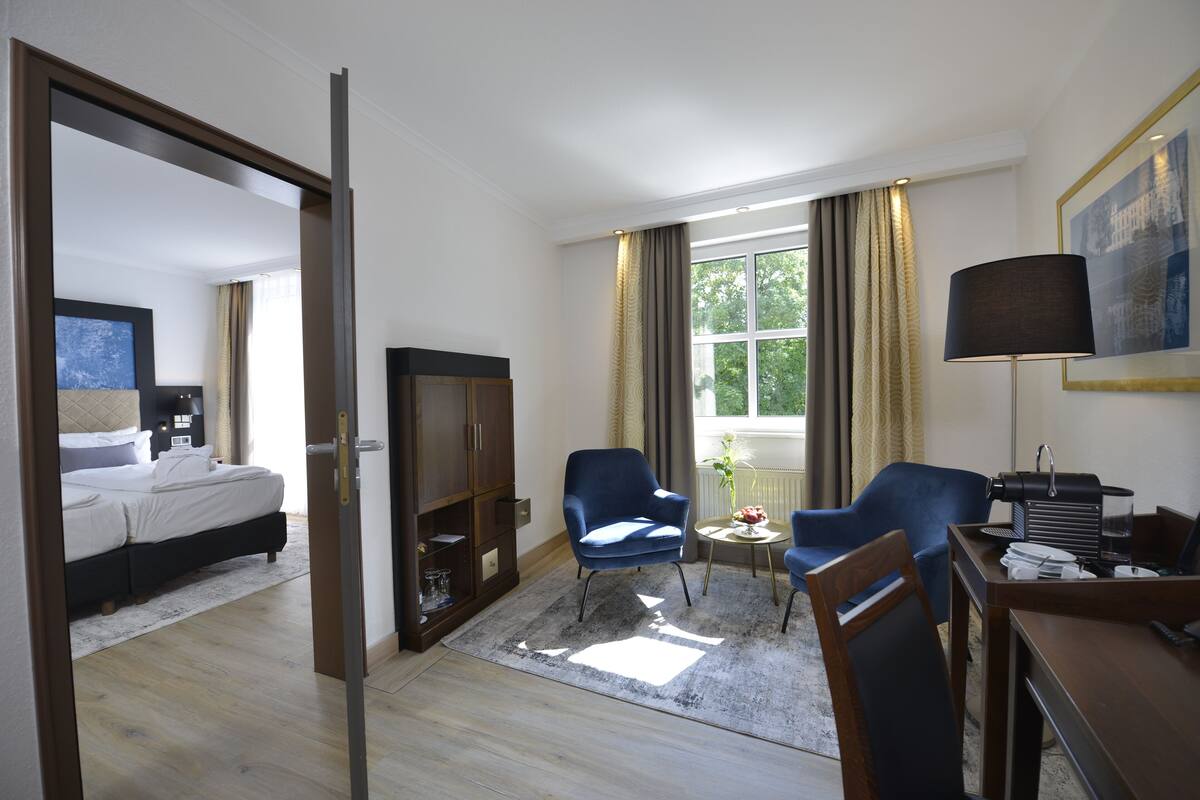 A hotel room with a bed, a desk, two chairs, a lamp, and a window with curtains.
