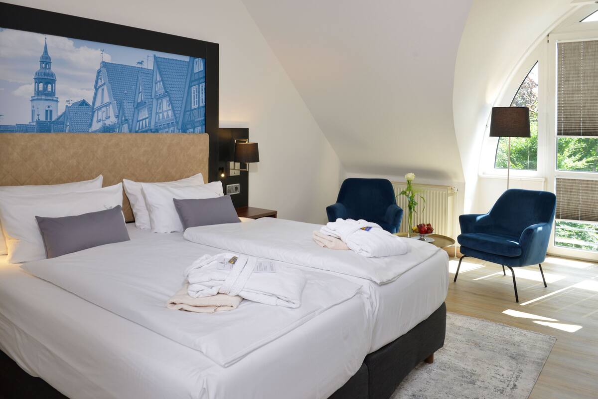 A hotel room with two beds, a large window, a blue chair, and a painting of buildings on the wall.