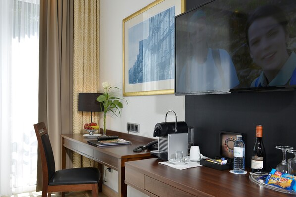 Habitación de hotel con escritorio, silla, televisor, un cuadro enmarcado y varios servicios, incluyendo una botella de agua, vino, aperitivos y un jarrón con flores.