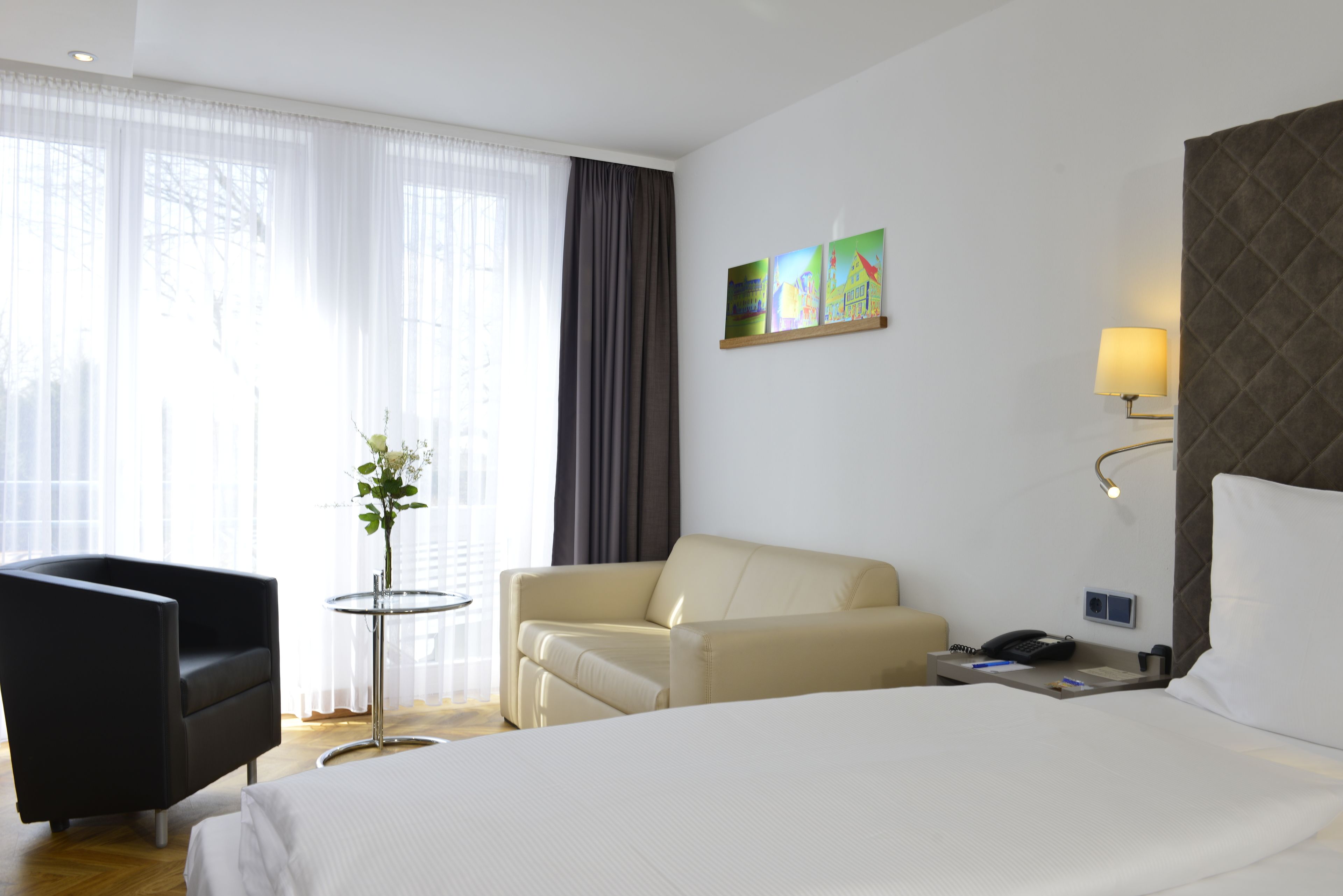 Une chambre d’hôtel avec un lit, un canapé, une chaise, une petite table avec une plante et des œuvres d’art accrochées au mur.