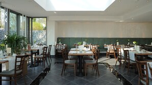 A modern restaurant with a high ceiling, large windows, and a green wall. Tables are set with chairs, and there are plants on the tables. - Hotel Caroline Mathilde (Celle)