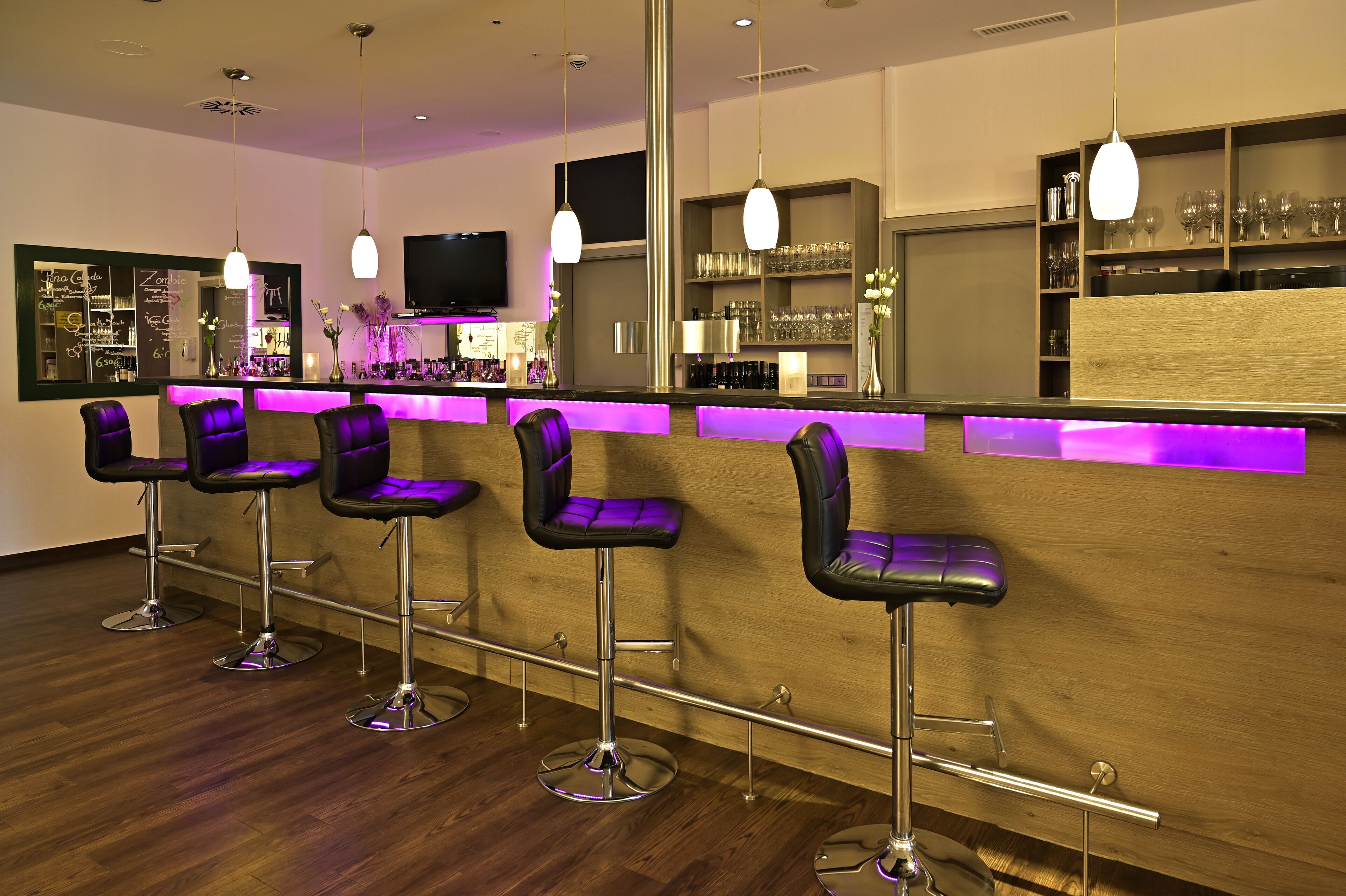 Zona de bar con cuatro taburetes, una barra iluminada de color púrpura y estantes con vasos y botellas.