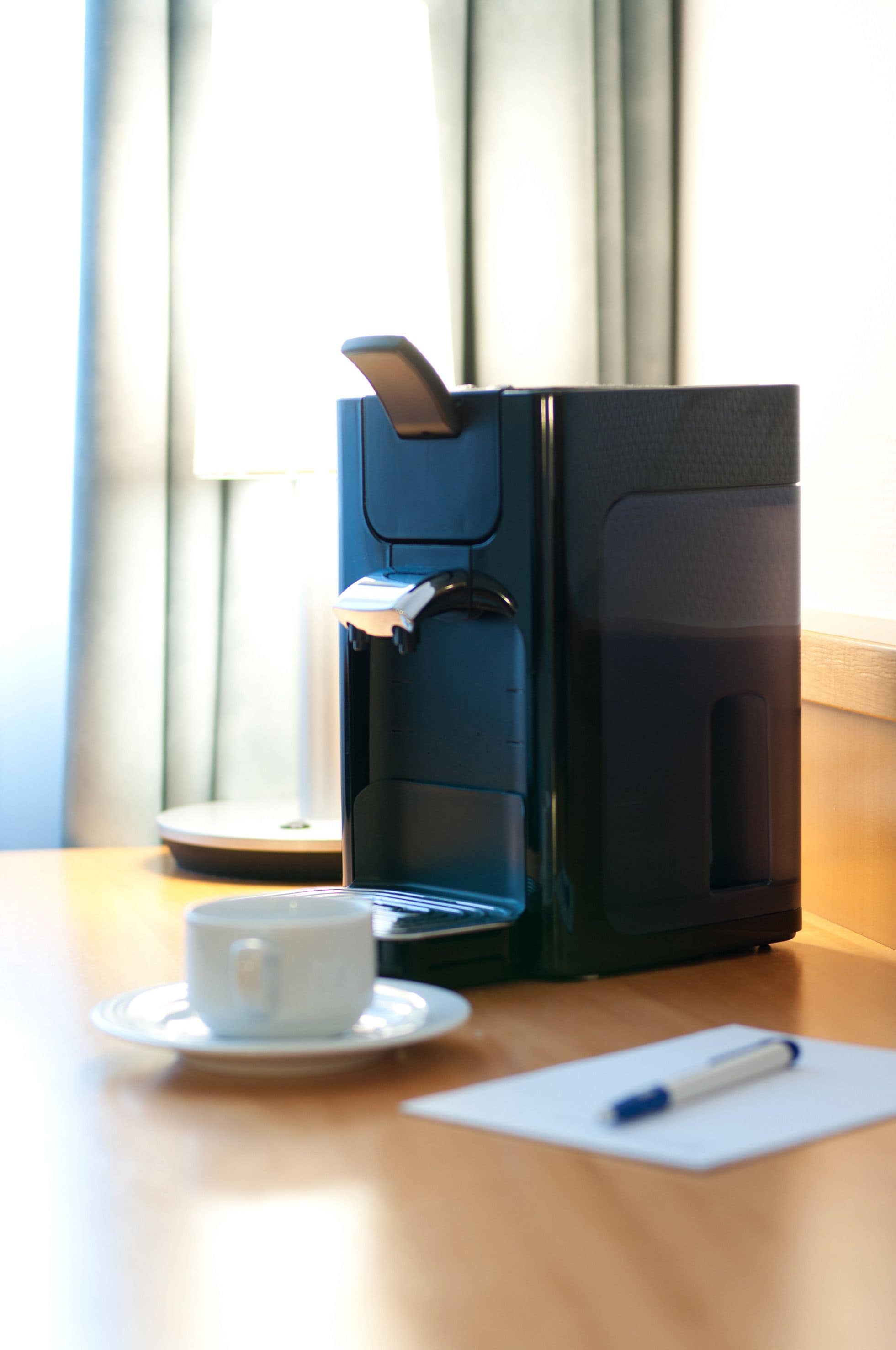 A coffee maker on a table with a cup and a pen.