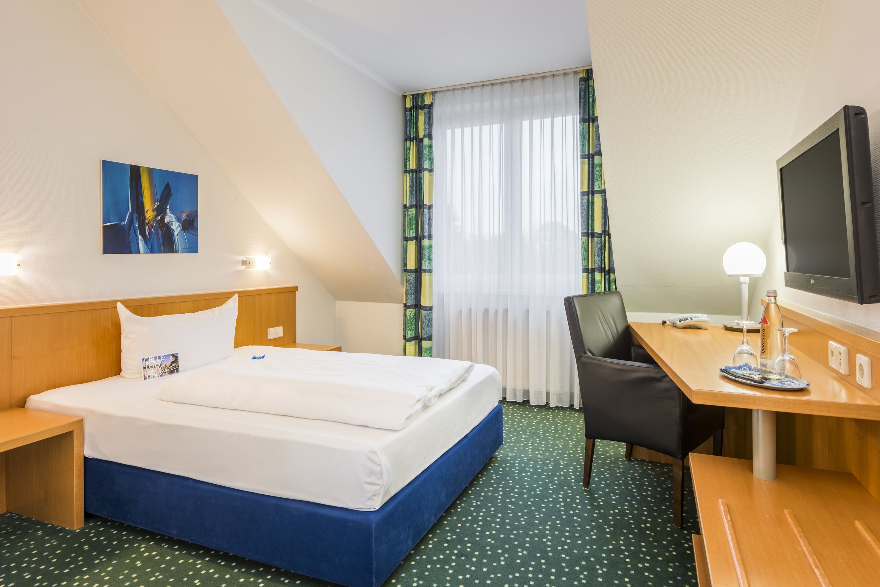 A hotel room with a bed, desk, chair, TV, and artwork on the wall.