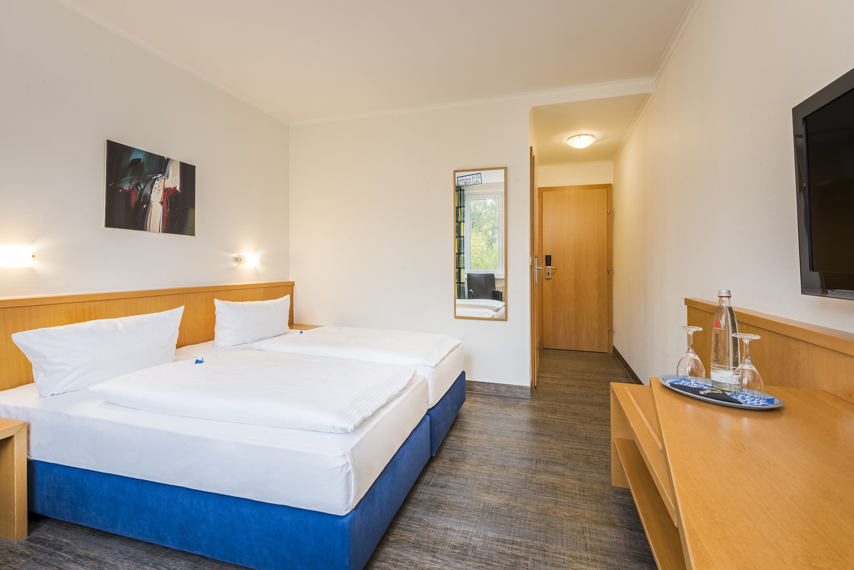 Et hotelværelse med en dobbeltseng, sengeborde, et skrivebord med stol, et spejl og et billede på væggen.