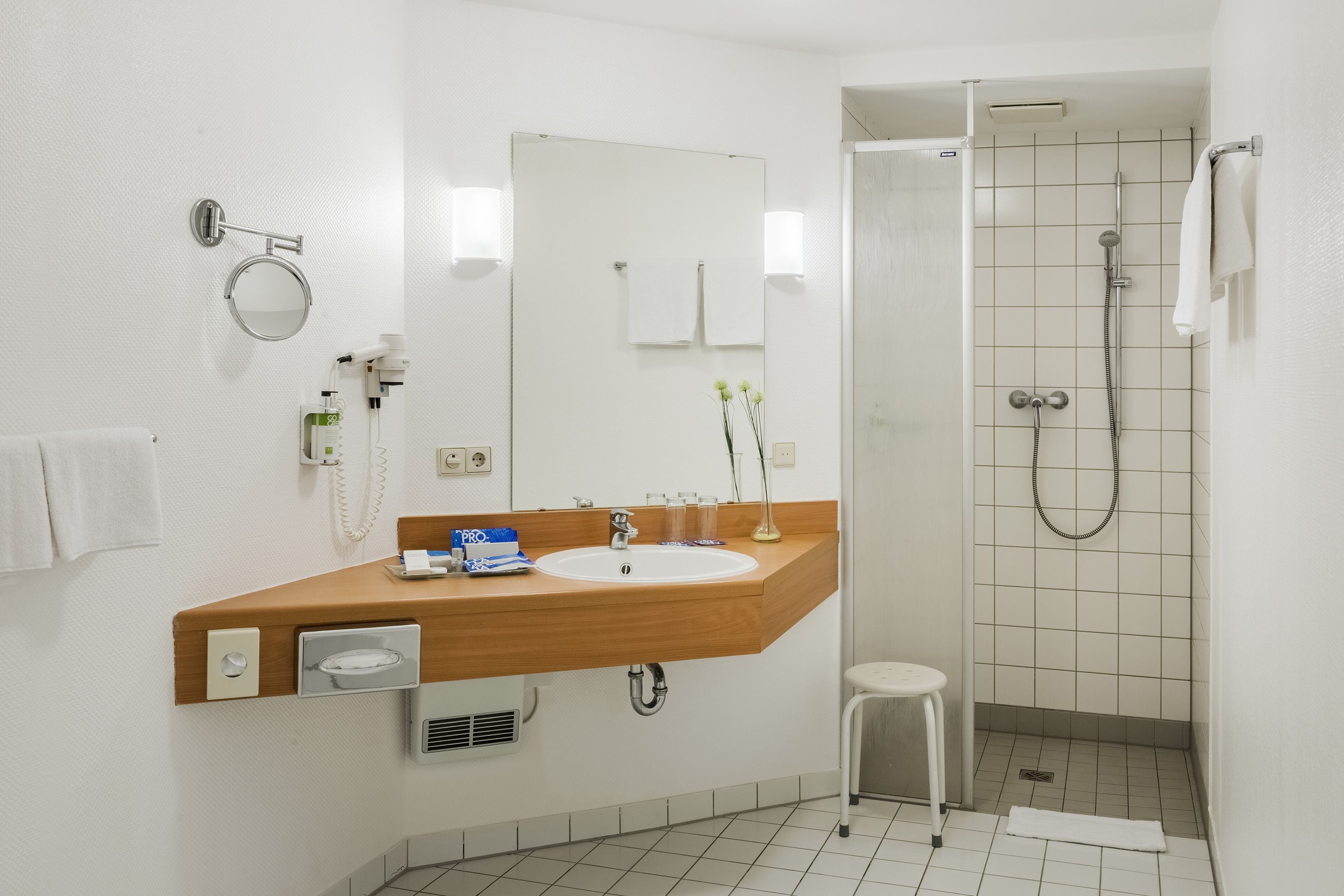 A modern bathroom with a wooden vanity, a sink, a mirror, a shower, and a small stool.