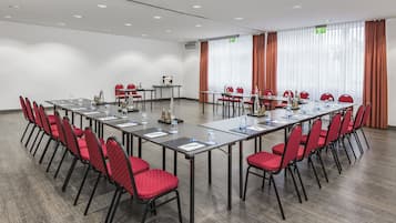 Un salón de conferencias con mesas largas, sillas rojas y cortinas blancas.