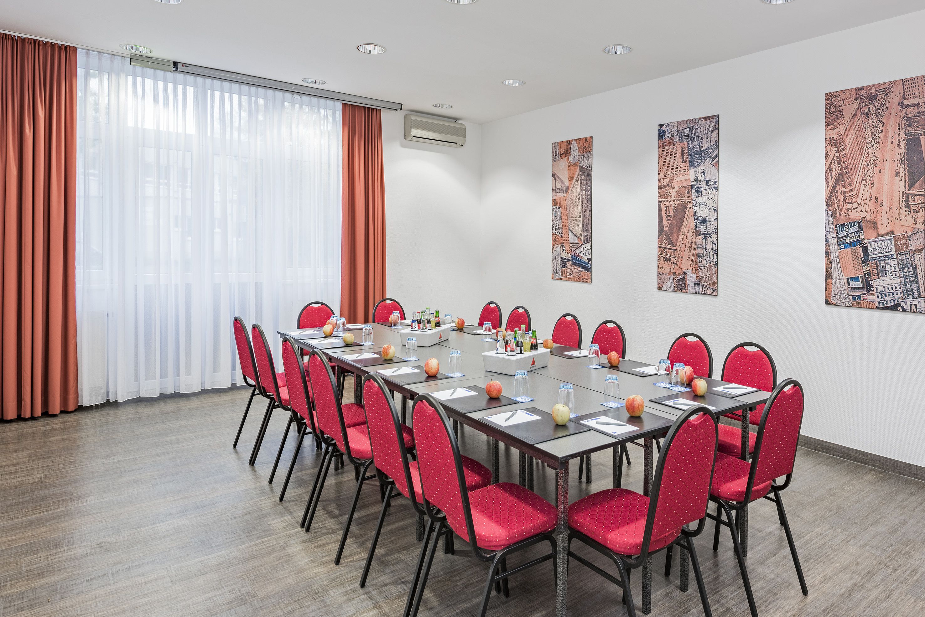 Een vergaderzaal met een lange tafel, rode stoelen en twee grote stadsgezichtschilderijen aan de muur.