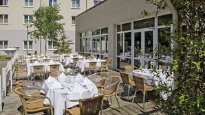 An outdoor dining area with wicker chairs and tables set up on a wooden deck, overlooking a building with large windows. - Hesse Hotel Celle (Celle)