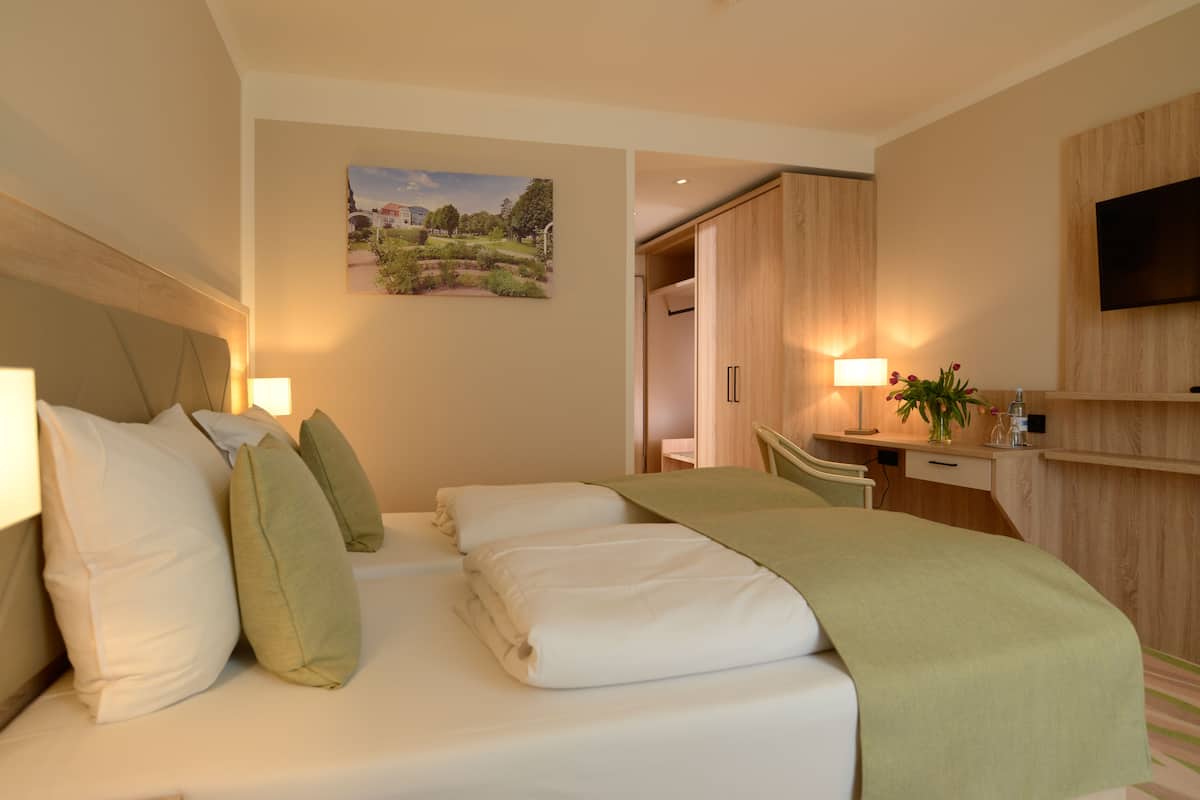 A hotel room with a bed, bedside lamps, a desk, a chair, a TV, and a painting on the wall.
