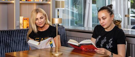 Two individuals seated at a wooden table in a modern room with bookshelves and a lamp.