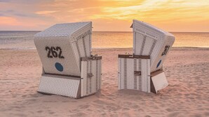 Two white wicker chairs with numbers on them, placed on a sandy beach with a sunset in the background. - TOP CountryLine Seehotel Großherzog Ostseebad Boltenhagen (Boltenhagen)