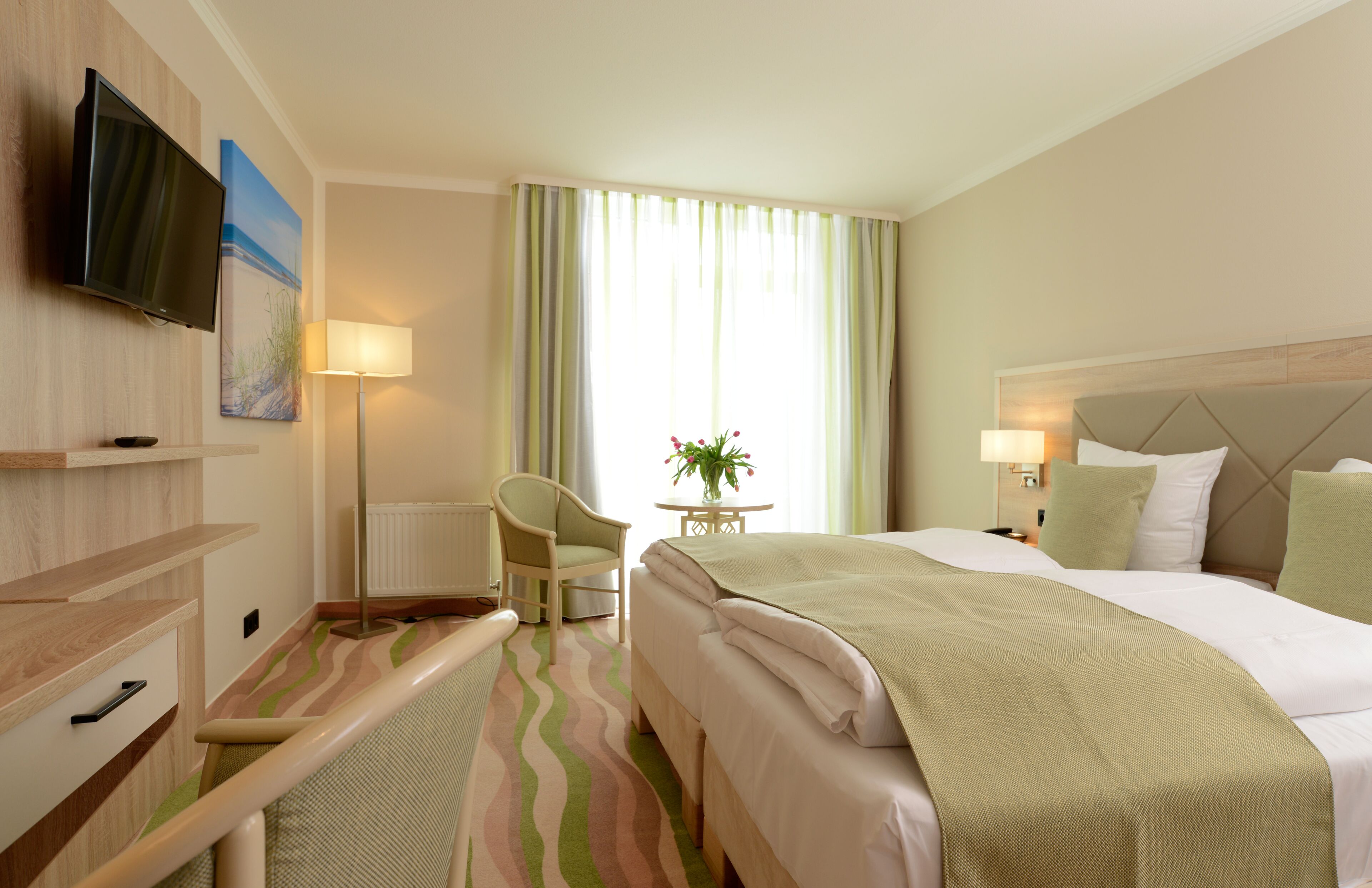 Ein Hotelzimmer mit Bett, Sessel, Tisch mit Pflanze, an der Wand befestigtem Fernseher und einem Fenster mit Vorhängen.