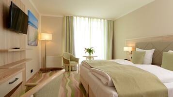 Ein Hotelzimmer mit Bett, Sessel, Tisch mit Pflanze, an der Wand befestigtem Fernseher und einem Fenster mit Vorhängen.