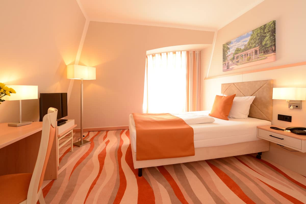 A hotel room with a bed, bedside table, lamp, and a painting on the wall.