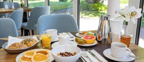 A breakfast table with a variety of food items, including pancakes, a bowl of cereal, fresh fruit, and a cup of tea.