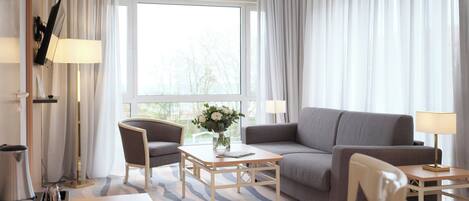 Habitación de hotel con sofá, sillón, mesa de centro y vistas a árboles a través de la ventana.