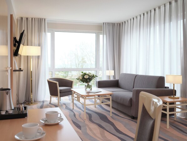 A hotel room with a sofa, armchair, coffee table, and a view of trees through the window.