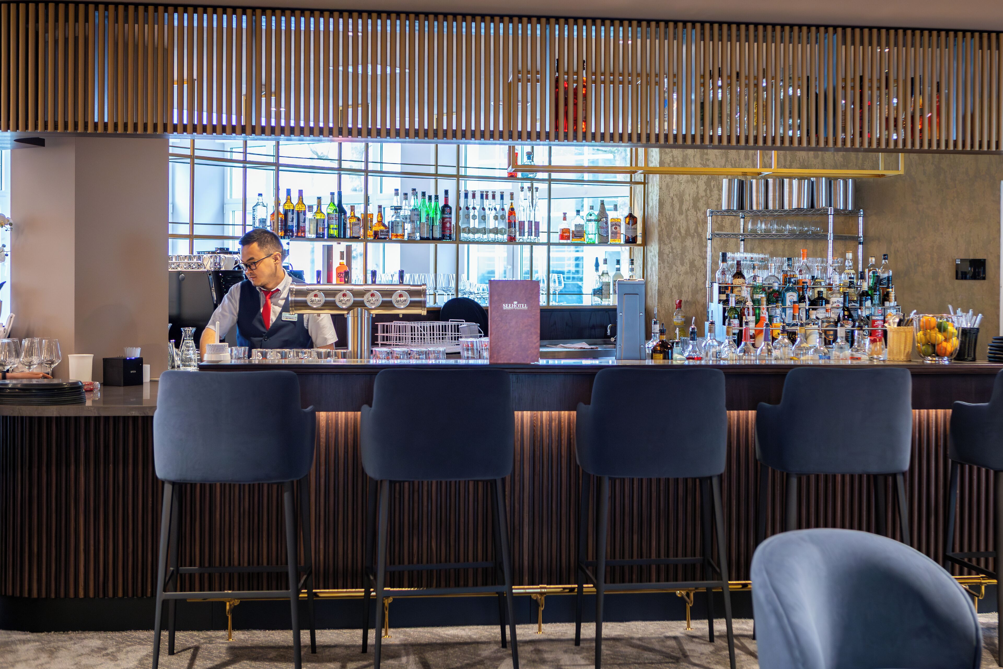 Zona de bar con un cantinero, taburetes y una variedad de botellas y vasos en estanterías.
