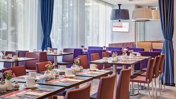 A hotel dining area with tables set for breakfast, purple and brown chairs, and large windows with blue curtains.