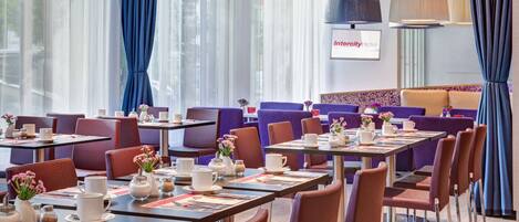 A hotel dining area with tables set for breakfast, purple and brown chairs, and large windows with blue curtains.