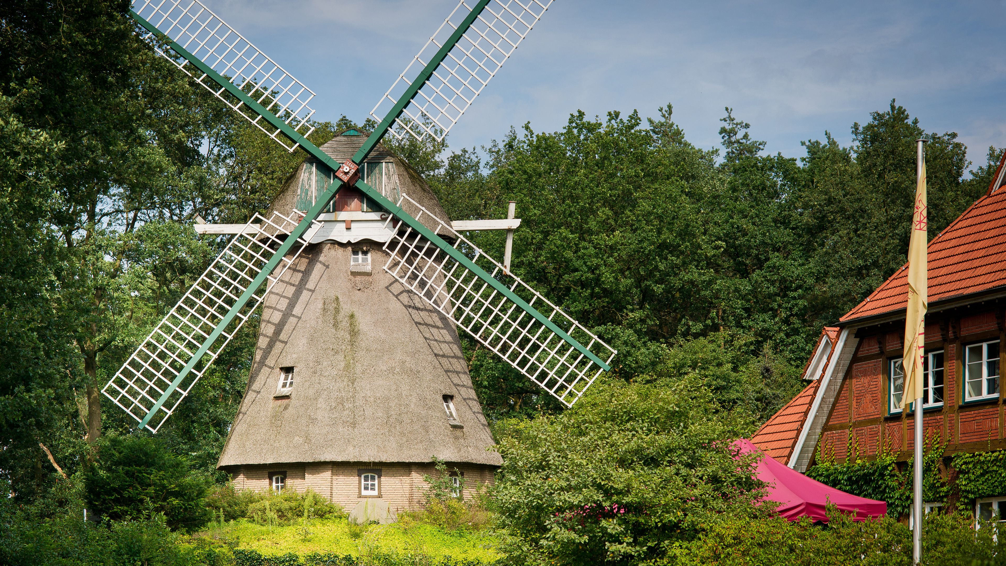 A traditional windmill with a thatched roof, surrounded by lush greenery and a red-roofed building.