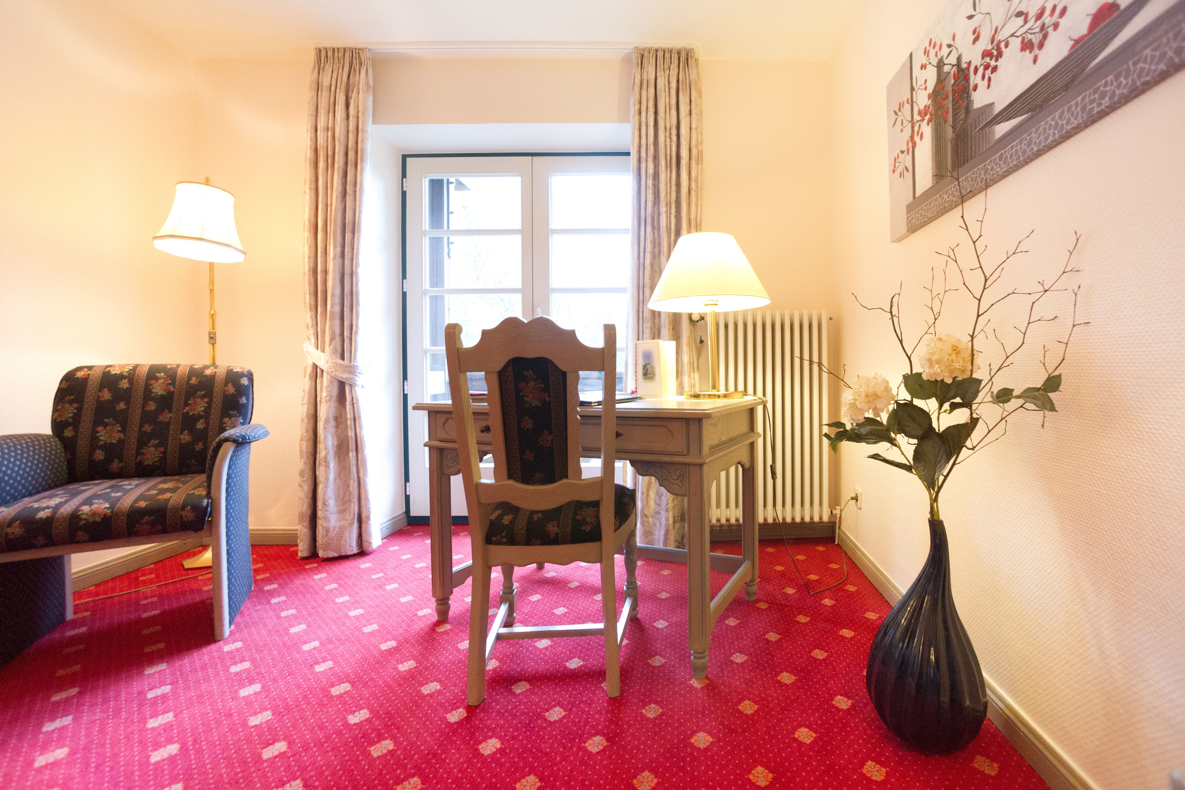 Una habitación con una silla de madera, un escritorio, una lámpara, un florero con flores y una alfombra estampada.
