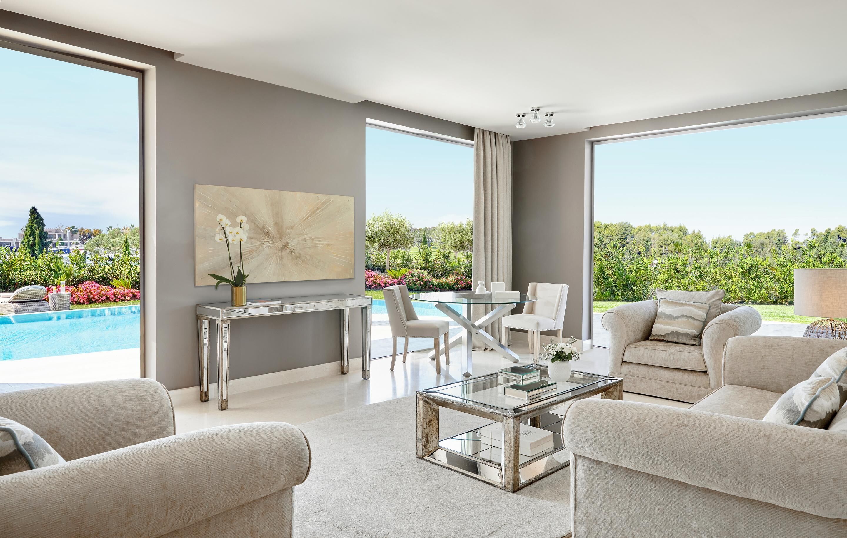 A modern living room with a glass coffee table, beige sofas, a dining area with a glass table, and a large window overlooking a pool and greenery.