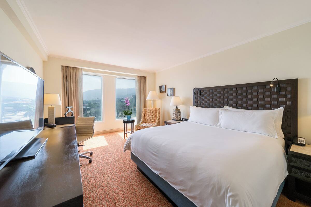 A hotel room with a large bed, a desk with a computer, a chair, and a view of the city through large windows.