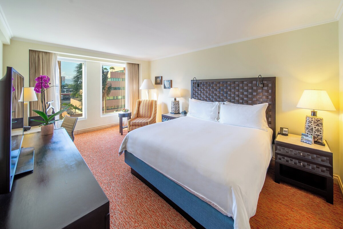 Ein Hotelzimmer mit einem großen Bett, einem Schreibtisch mit Computer, einem Stuhl und einem Fenster mit Vorhängen.
