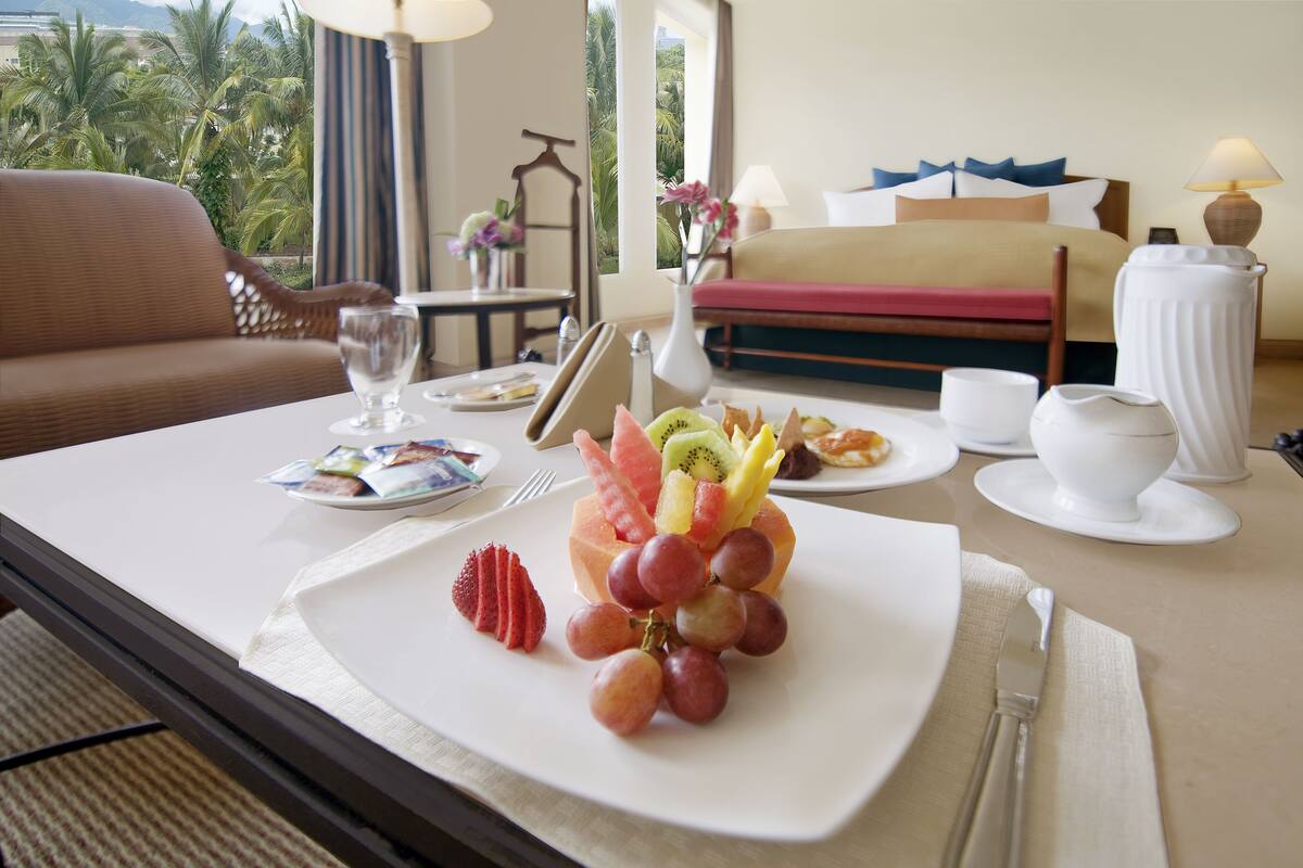 A hotel room with a dining table set for breakfast, including a fruit platter, a plate of pastries, and a cup of coffee.