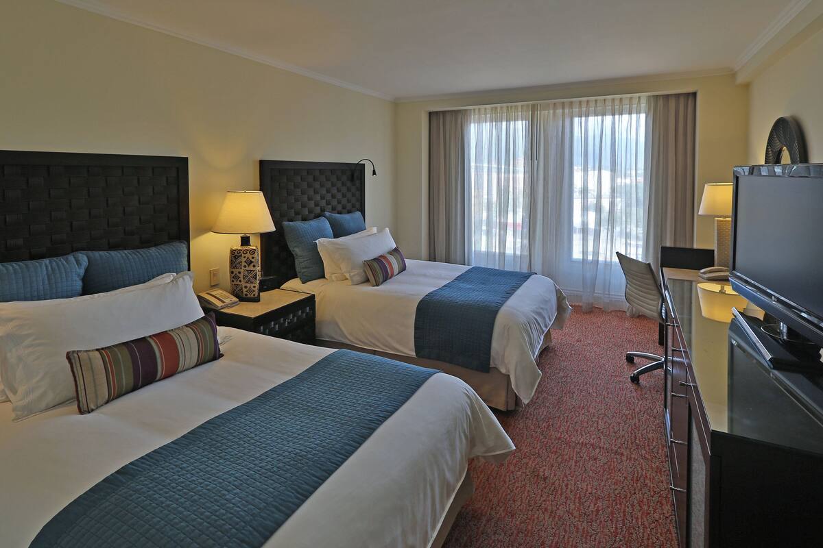 Une chambre d’hôtel avec deux lits, un bureau avec une télévision et une grande fenêtre avec des rideaux.