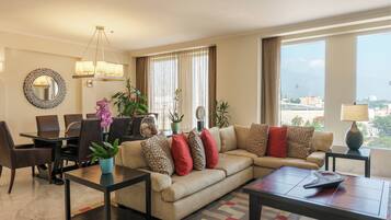 A modern living room with a sofa, coffee table, and dining area. There is a large window with curtains, a chandelier, and a view of the city.