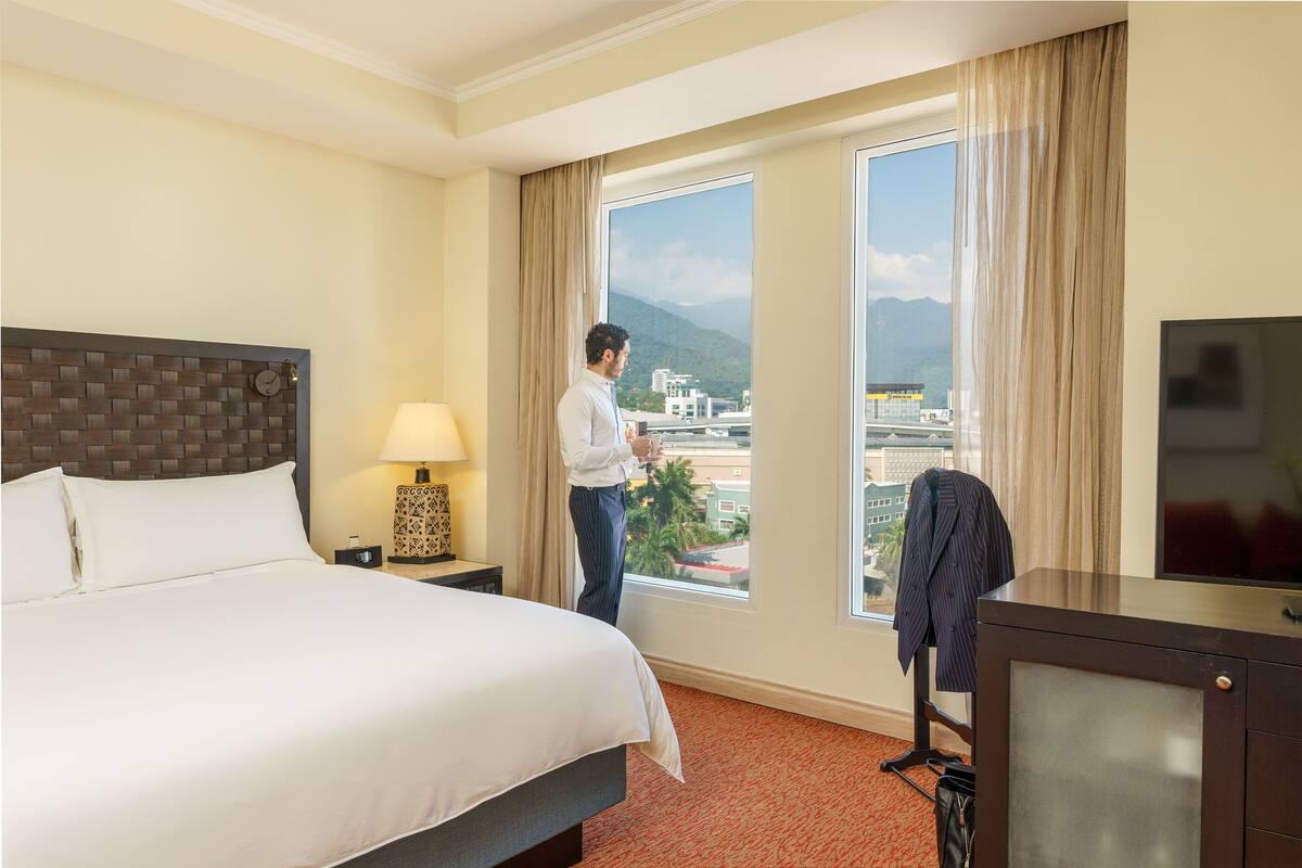 A hotel room with a large bed, a desk with a TV, a window with a view of the city and mountains, and a lamp on the nightstand.