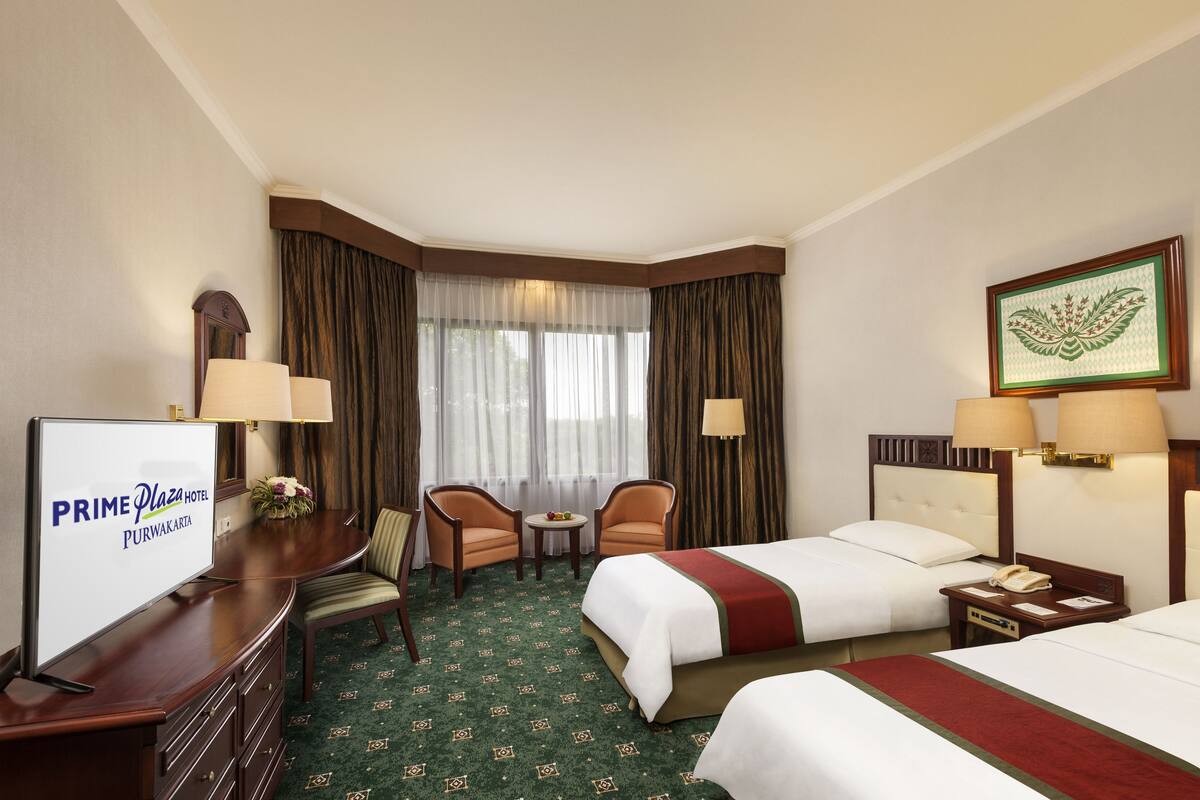 A hotel room with two beds, a desk with a TV, two chairs, a small table, and a window with curtains.
