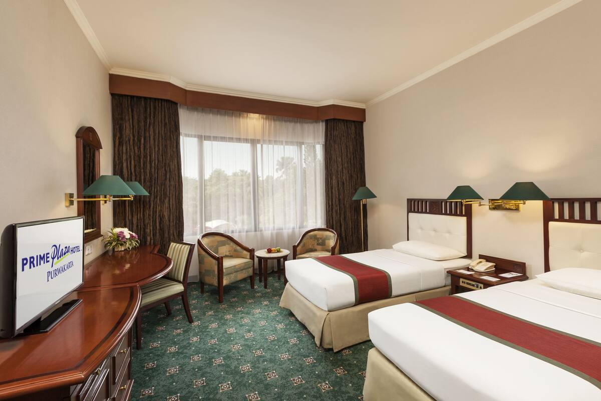 A hotel room with two beds, a desk with a television, a small table with a vase of flowers, and a window with curtains.
